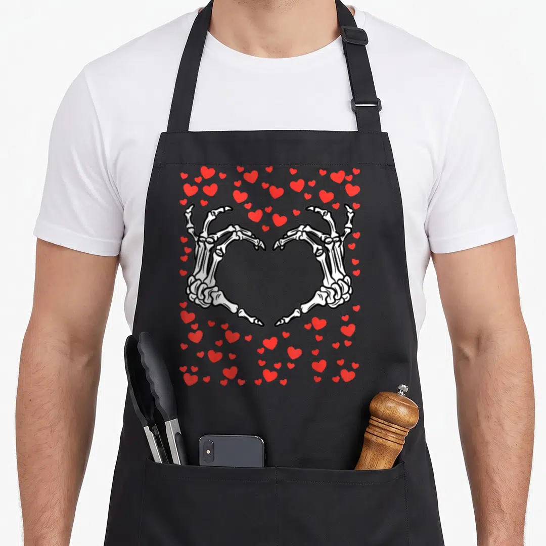 Valentine skeleton apron - Two white skeleton hands forming a heart shape surrounded by scattered red hearts in cartoon — shown on a black cotton canvas apron against a white background