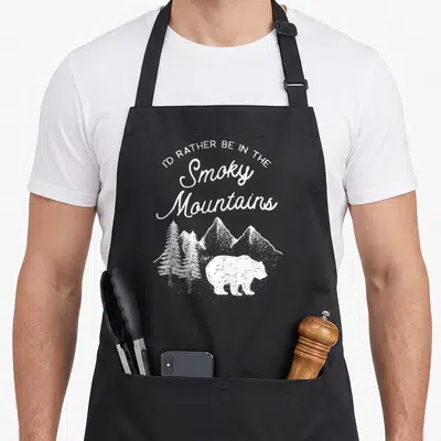 Smoky gatlinburg apron - Distressed white bear walking near pine trees under arched text reading I'd rather be — shown on a black cotton canvas apron against a white background