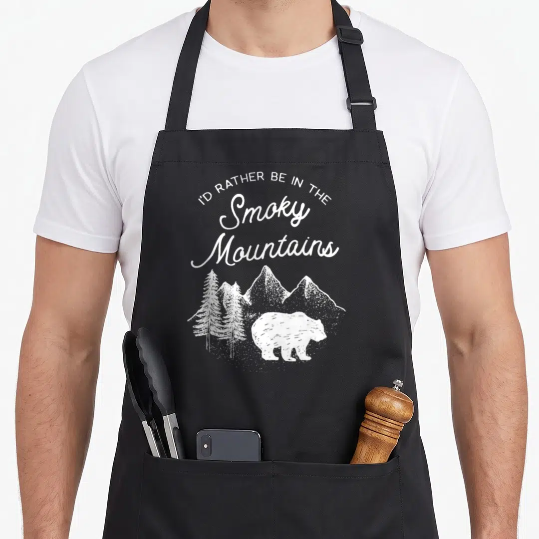 Smoky gatlinburg apron - Distressed white bear walking near pine trees under arched text reading I'd rather be — shown on a black cotton canvas apron against a white background