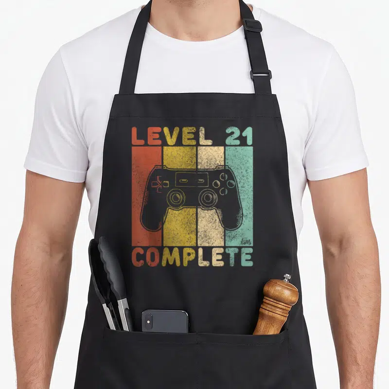 Complete gaming aprons - Grey game controller over four vertical red yellow beige and teal stripes with retro text — shown on a black cotton canvas apron against a white background