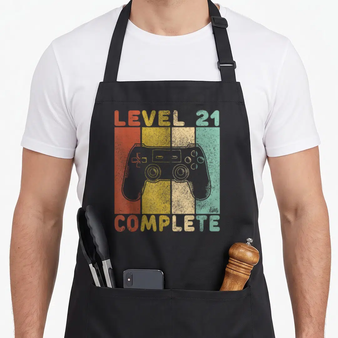 Complete gaming aprons - Grey game controller over four vertical red yellow beige and teal stripes with retro text — shown on a black cotton canvas apron against a white background