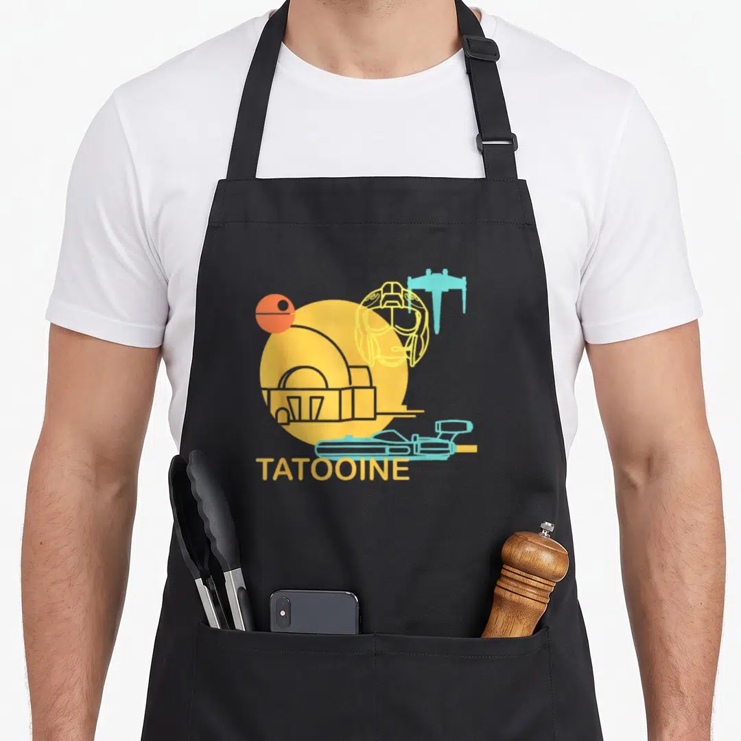 Drawing new hope apron - Yellow circle with building silhouette, blue ship, orange sphere, and TATOOINE text — shown on a black cotton canvas apron against a white background