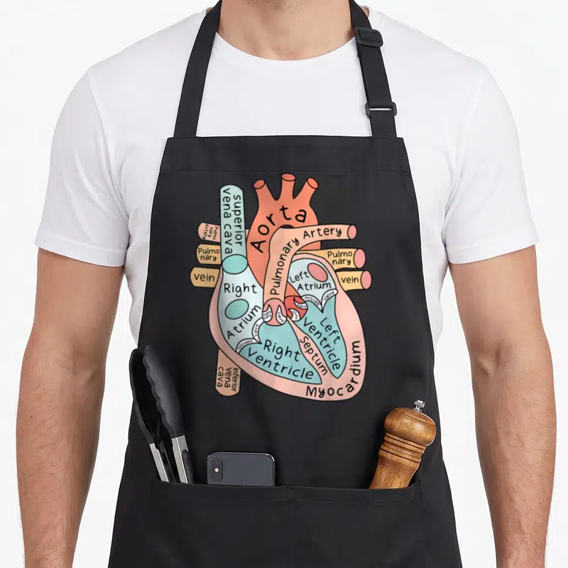 Anatomical nursing student apron - Cartoon heart diagram with labels like Aorta, valves, and ventricles in pink, blue — shown on a black cotton canvas apron against a white background