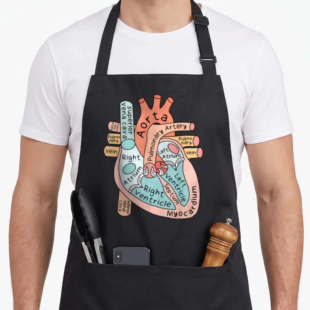 Anatomical nursing student apron - Cartoon heart diagram with labels like Aorta, valves, and ventricles in pink, blue — shown on a black cotton canvas apron against a white background
