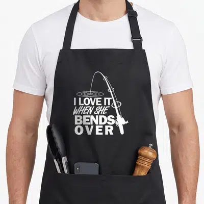 Bend fishing aprons - White bent fishing rod with ripples and stacked text I LOVE IT WHEN SHE BENDS — shown on a black cotton canvas apron against a white background