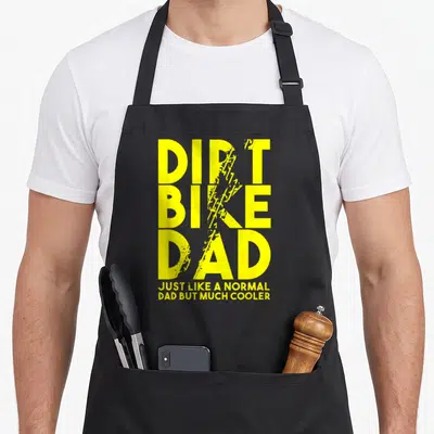 Dirt father day apron - Yellow distressed text DIRT BIKE DAD with tire track overlays and cooler dad quote — shown on a black cotton canvas apron against a white background
