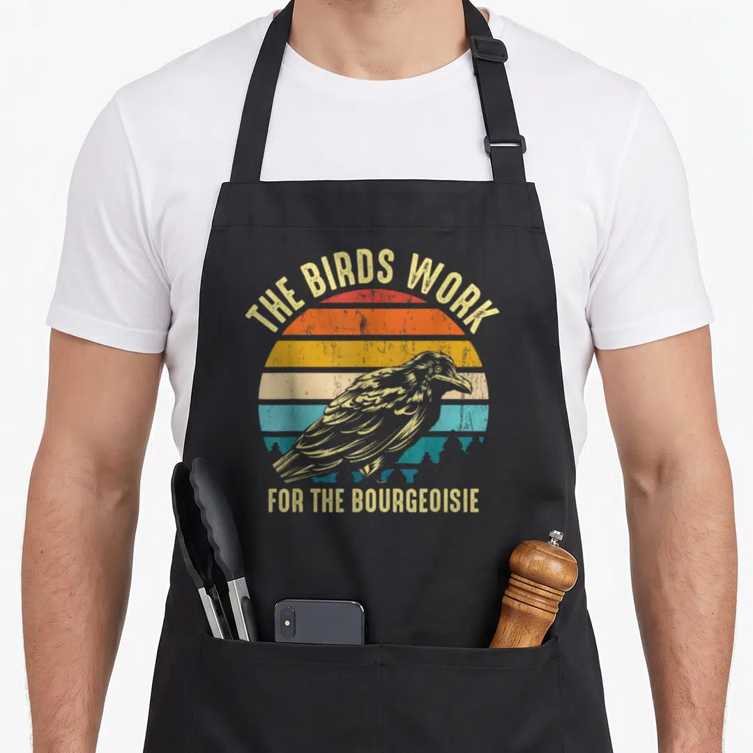 Bourgeoisie birds apron - Retro crow illustration over beige red orange and blue striped sun with sarcastic text — shown on a black cotton canvas apron against a white background