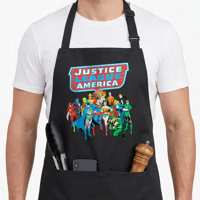 Justice group shot apron - Justice League of America shield banner with tiered superhero team in red blue yellow — shown on a black cotton canvas apron against a white background
