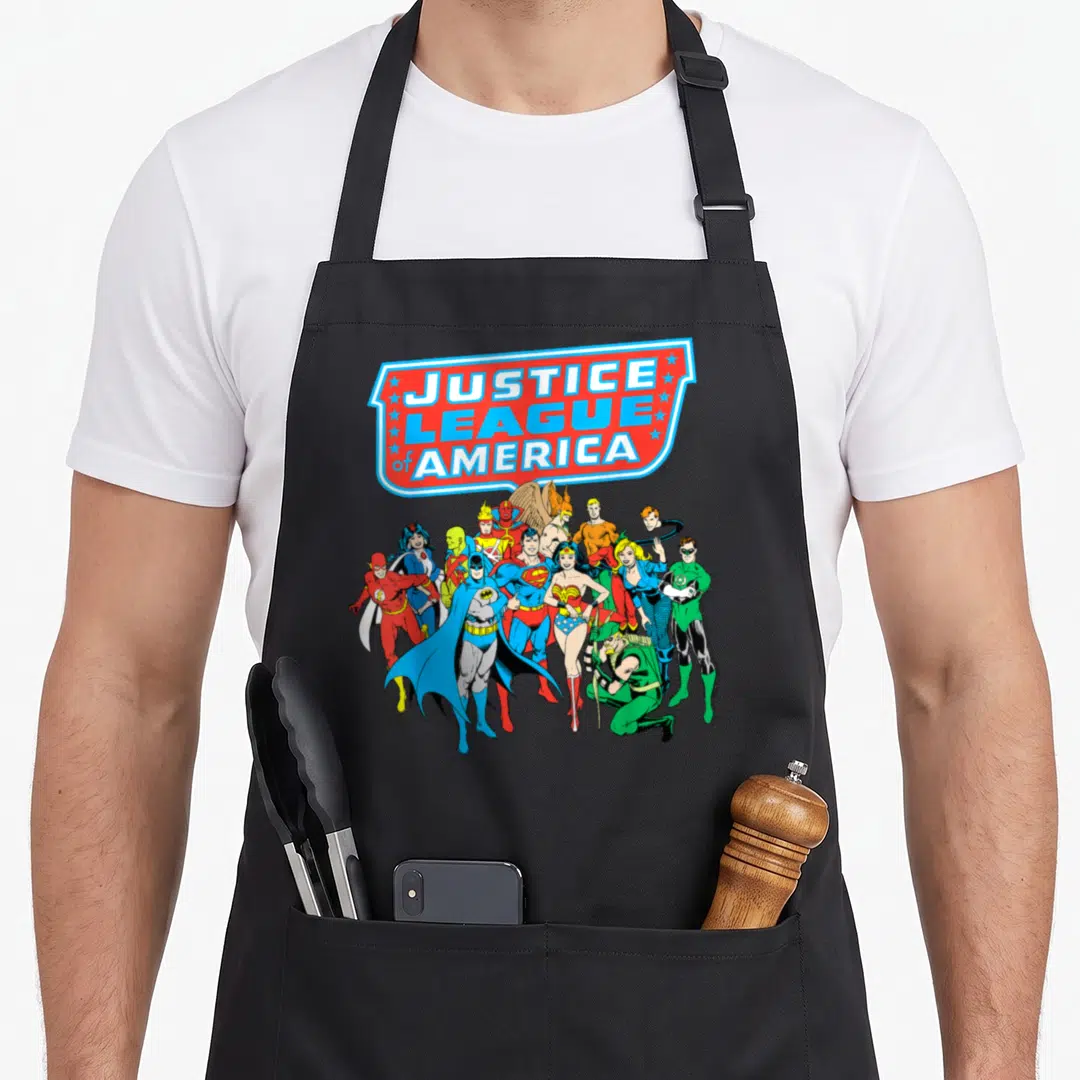 Justice group shot apron - Justice League of America shield banner with tiered superhero team in red blue yellow — shown on a black cotton canvas apron against a white background