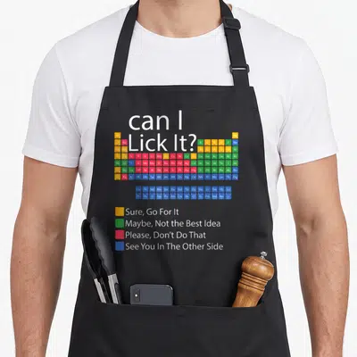 Periodic chemistry apron - Can I Lick It text above colorful periodic table with safety legend squares — shown on a black cotton canvas apron against a white background