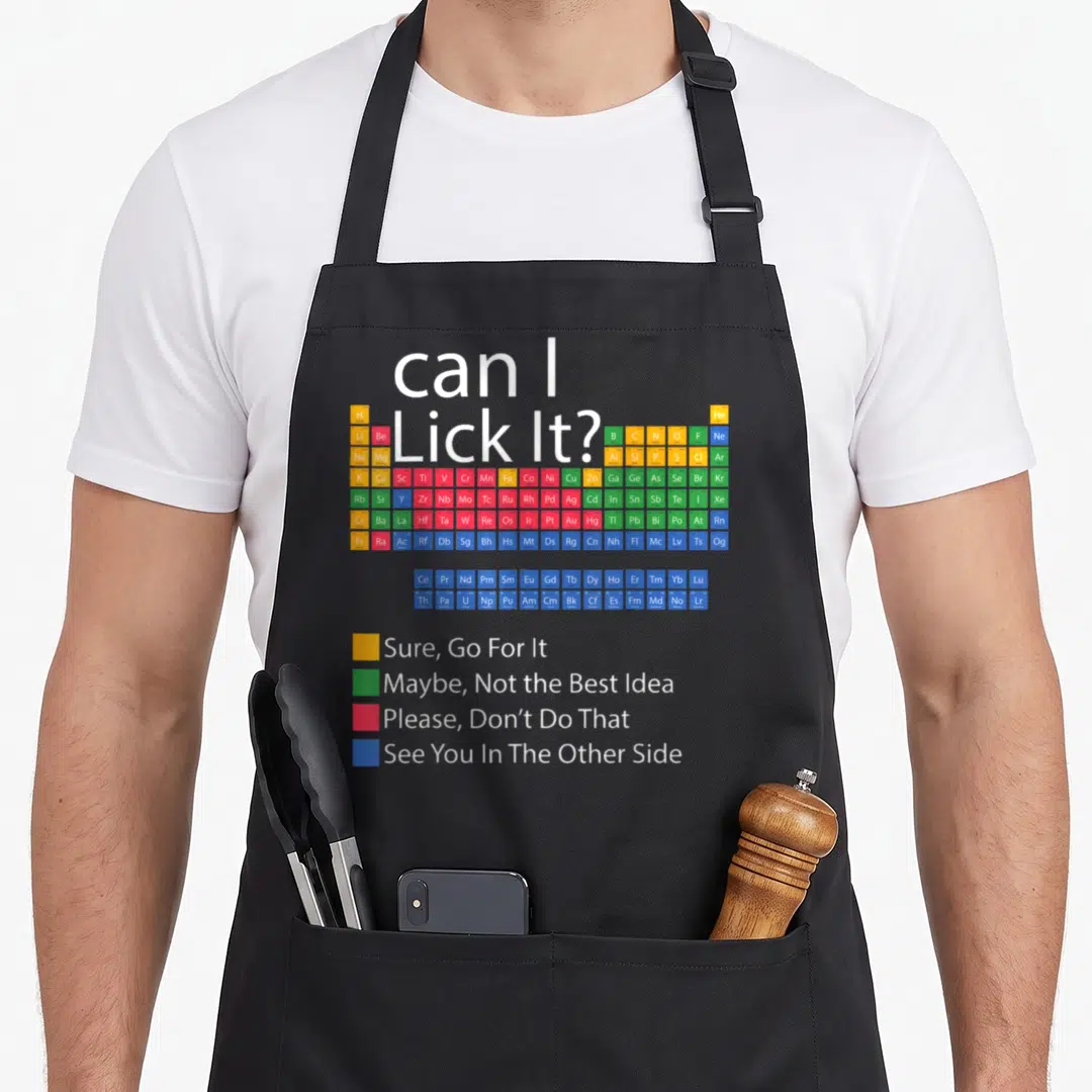 Periodic chemistry apron - Can I Lick It text above colorful periodic table with safety legend squares — shown on a black cotton canvas apron against a white background