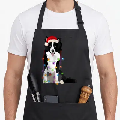 Christmas border collie apron - Cartoon border collie in red santa hat wrapped in multicolored christmas lights — shown on a black cotton canvas apron against a white background
