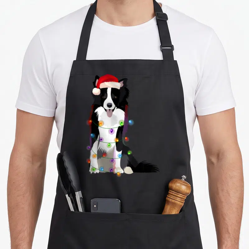 Christmas border collie apron - Cartoon border collie in red santa hat wrapped in multicolored christmas lights — shown on a black cotton canvas apron against a white background