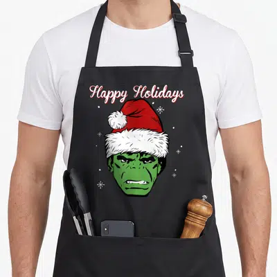 Holiday santa aprons - Green Hulk head wearing red Santa hat below arched Happy Holidays text surrounded by snowflakes — shown on a black cotton canvas apron against a white background