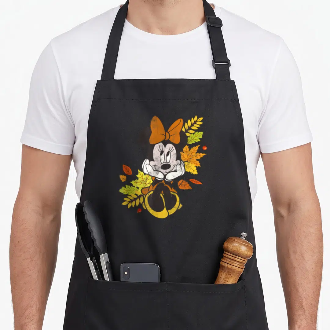 Autumn friends apron - Minnie Mouse sitting with chin on hands surrounded by orange and yellow autumn leaves and small — shown on a black cotton canvas apron against a white background