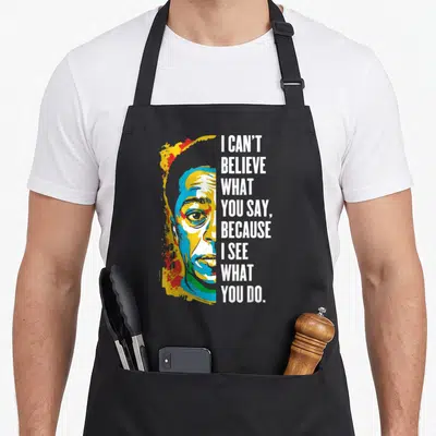 Jame black history apron - Pop art portrait of James Baldwin next to stacked white text I can't believe what you say — shown on a black cotton canvas apron against a white background