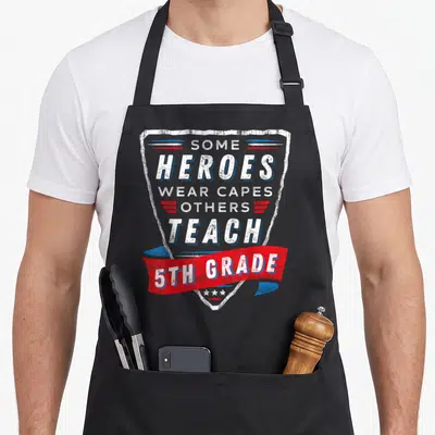 Heroe art teacher aprons - Distressed shield with SOME HEROES WEAR CAPES OTHERS TEACH 5TH GRADE text and red ribbon — shown on a black cotton canvas apron against a white background
