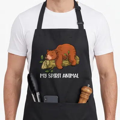 Vintage grizzly bear apron - Cartoon brown bear sleeping on tan log with green leaves and white text reading My Spirit — shown on a black cotton canvas apron against a white background