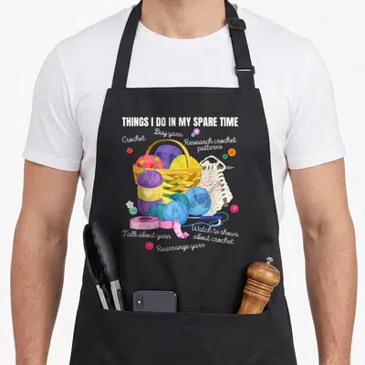 Spare craft apron - Cartoon basket of purple blue and pink yarn surrounded by crochet hooks and buttons with white text — shown on a black cotton canvas apron against a white background