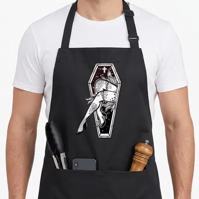Zombie tattoo apron - Tattooed legs dangling from coffin with cross and stars in line art — shown on a black cotton canvas apron against a white background