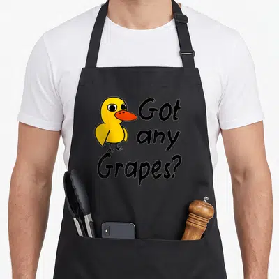 Grape song duck apron - Cartoon yellow duck next to stacked text Got any Grapes? in black and orange — shown on a black cotton canvas apron against a white background