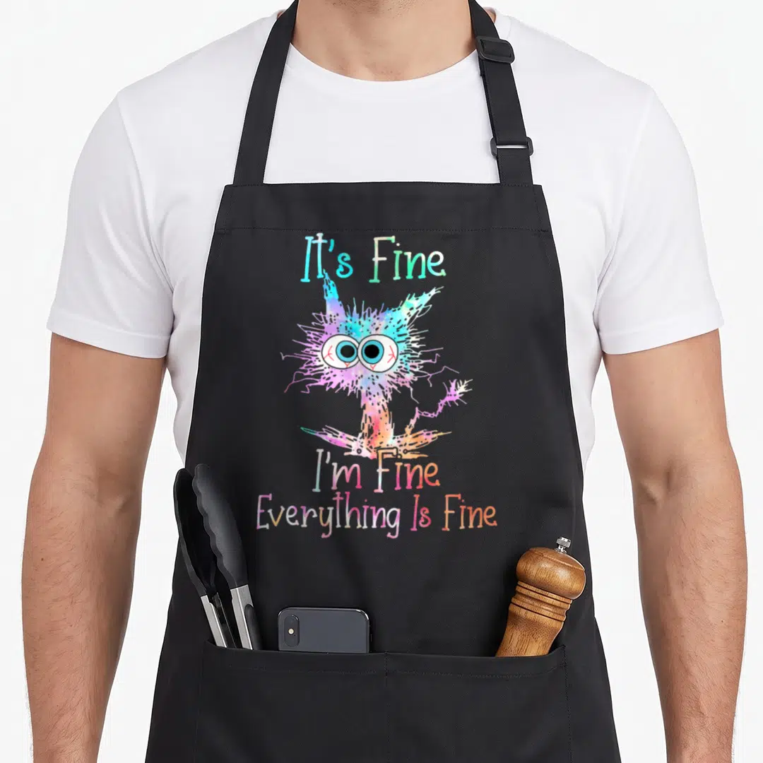 Fine tie-dye apron - Cartoon multicolored monster with It's Fine I'm Fine Everything Is Fine text — shown on a black cotton canvas apron against a white background