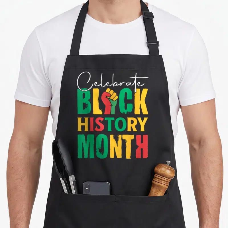 History celebrate apron - Stacked text Celebrate Black History Month with red raised fist replacing A in Black — shown on a black cotton canvas apron against a white background