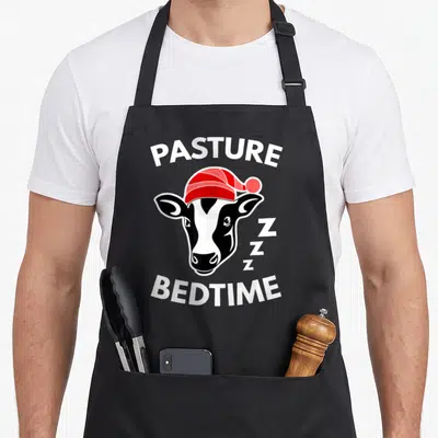 Pasture cow apron - Cartoon cow head in red nightcap with PASTURE BEDTIME text and three Zs — shown on a black cotton canvas apron against a white background