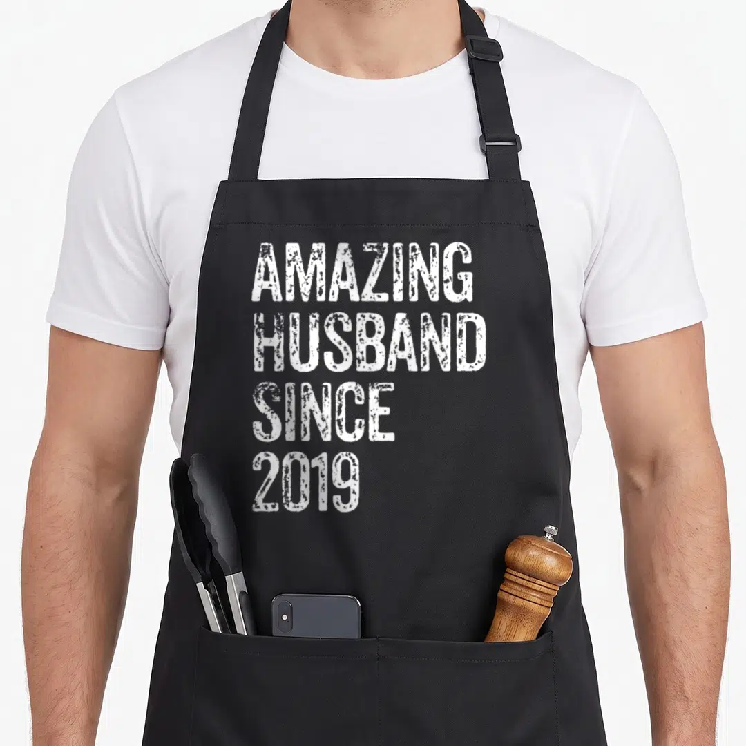 Amazing wedding apron - White distressed text reading AMAZING HUSBAND SINCE 2019 stacked vertically — shown on a black cotton canvas apron against a white background