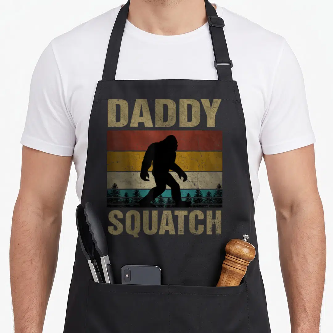 Daddy yeti apron - Retro bigfoot silhouette walking on red orange teal stripes with DADDY SQUATCH text — shown on a black cotton canvas apron against a white background