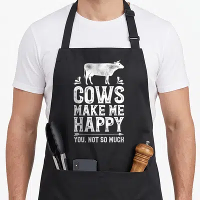 Much farmer apron - Distressed white cow standing above stacked text reading COWS MAKE ME HAPPY YOU NOT SO MUCH — shown on a black cotton canvas apron against a white background