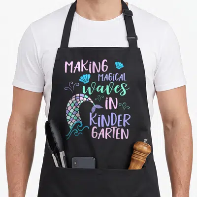 Kindergarten making an apron - Mermaid tail with pink purple mint scales and text MAKING MAGICAL Waves IN KINDER GARTEN — shown on a black cotton canvas apron against a white background