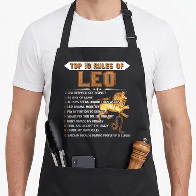 Leo zodiac birthday apron - Top 10 Rules of Leo text with leaping lion and zodiac symbol in orange and yellow — shown on a black cotton canvas apron against a white background