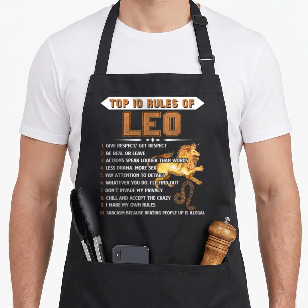 Leo zodiac birthday apron - Top 10 Rules of Leo text with leaping lion and zodiac symbol in orange and yellow — shown on a black cotton canvas apron against a white background