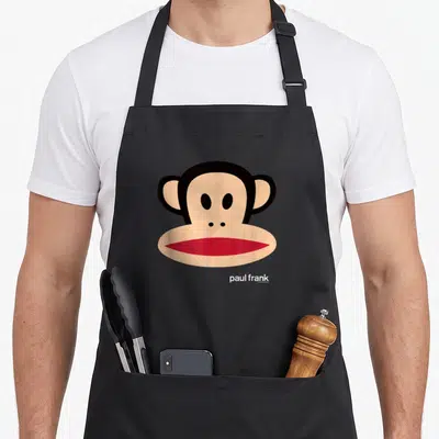Paul face apron - Cartoon monkey head in beige and black with red mouth and white text below — shown on a black cotton canvas apron against a white background