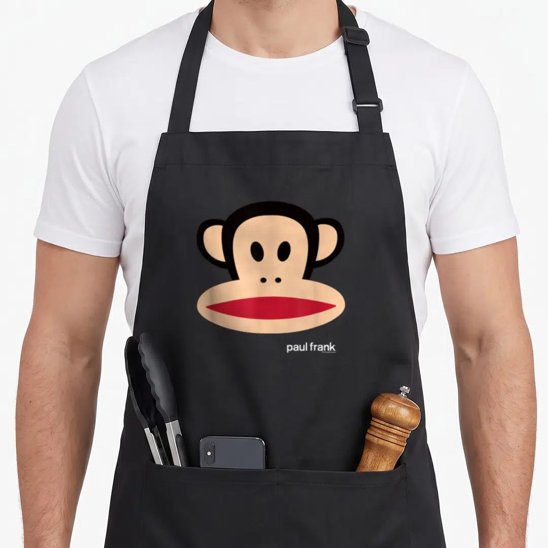 Paul face apron - Cartoon monkey head in beige and black with red mouth and white text below — shown on a black cotton canvas apron against a white background