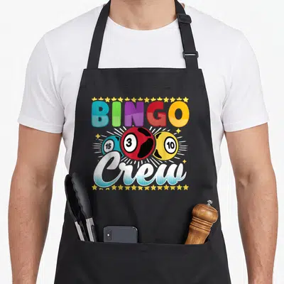 Crew bingo apron - BINGO Crew text above three numbered balls framed by yellow stars — shown on a black cotton canvas apron against a white background