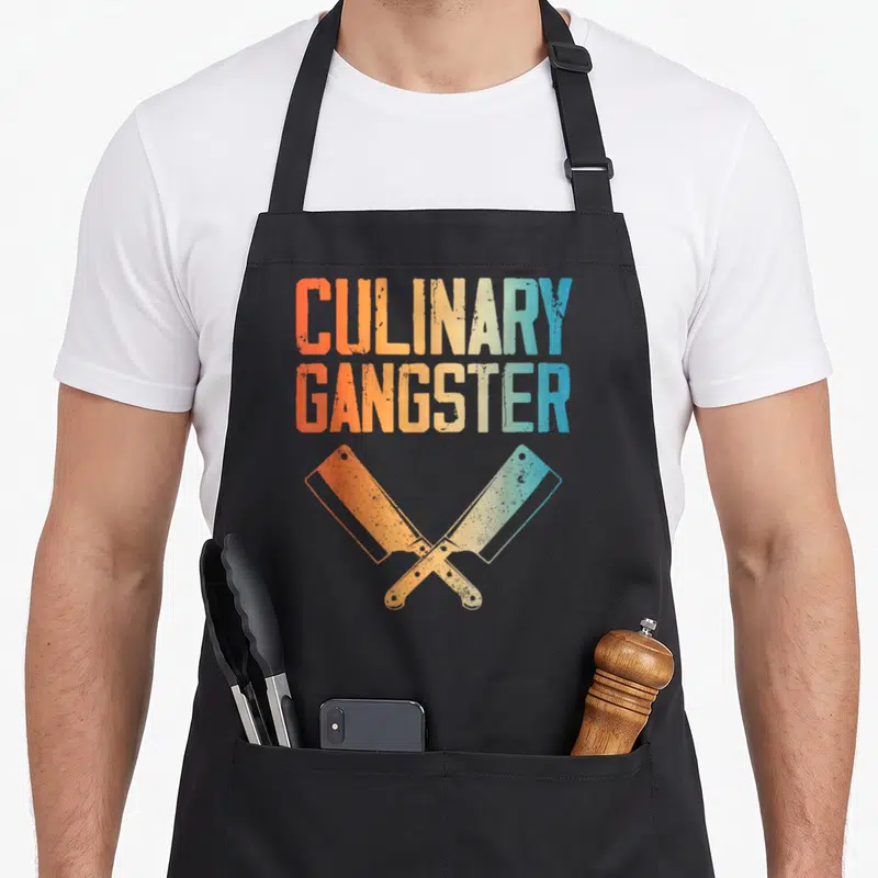 Sous culinary aprons - Crossed cleavers in orange yellow and blue below distressed CULINARY GANGSTER text — shown on a black cotton canvas apron against a white background
