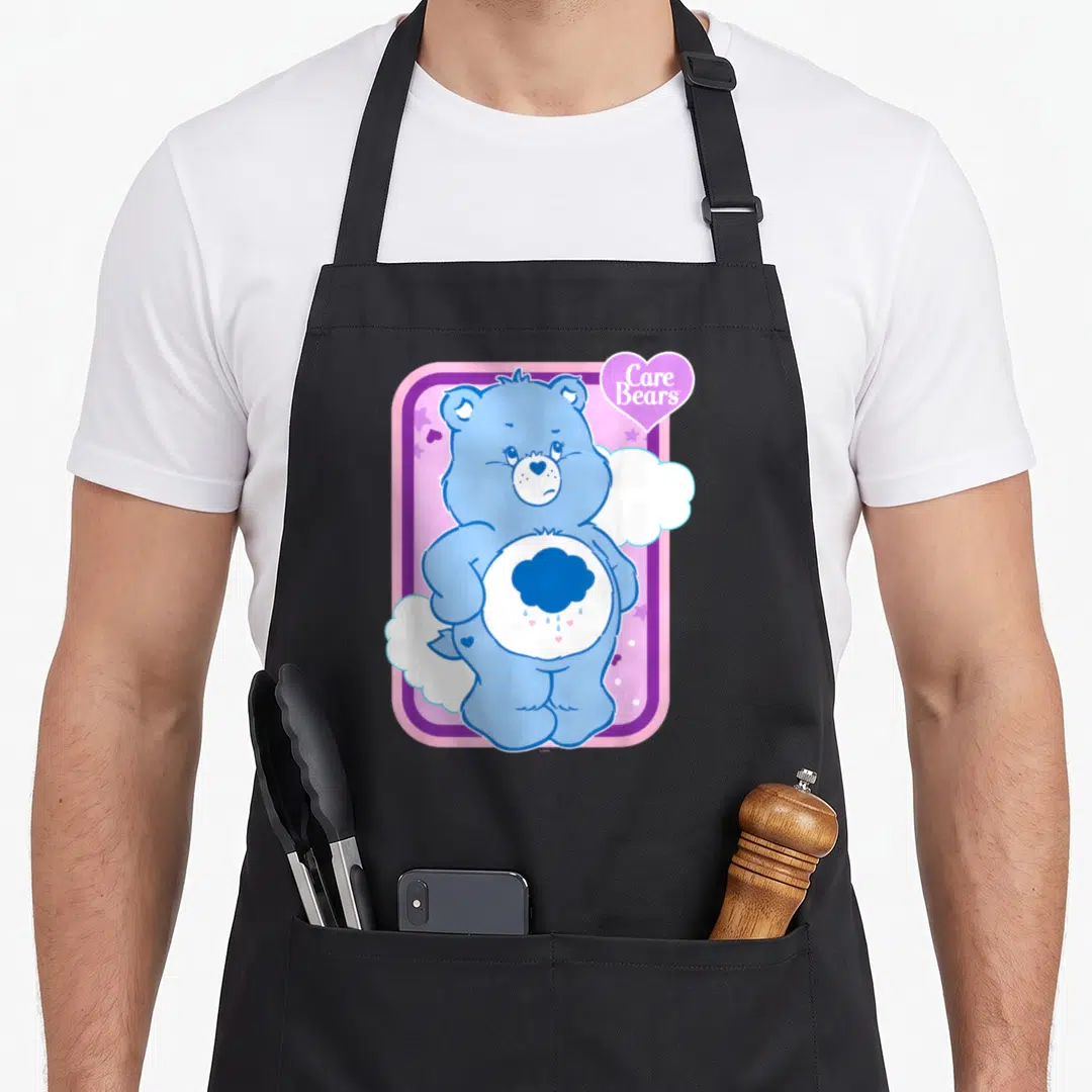 Grumpy care apron - Blue bear with rain cloud belly badge inside pink and purple frame with Care Bears text — shown on a black cotton canvas apron against a white background