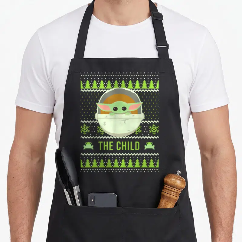 Funny christmas sweater apron - Grogu in pod with THE CHILD text and frog icons on grey green ugly sweater pattern — shown on a black cotton canvas apron against a white background