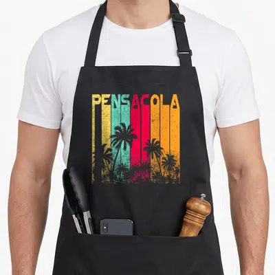 Pensacola holiday apron - PENSACOLA text over yellow teal red orange stripes with grey palm tree silhouettes — shown on a black cotton canvas apron against a white background
