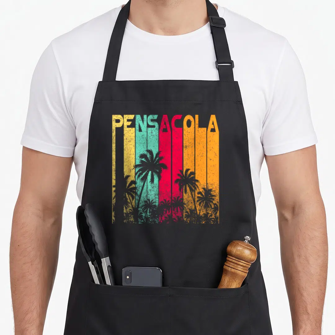 Pensacola holiday apron - PENSACOLA text over yellow teal red orange stripes with grey palm tree silhouettes — shown on a black cotton canvas apron against a white background
