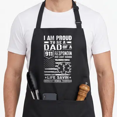 Proud emt apron - White distressed text I AM PROUD TO BE A DAD OF A 911 RESPONDIN over flag and Star of Life — shown on a black cotton canvas apron against a white background
