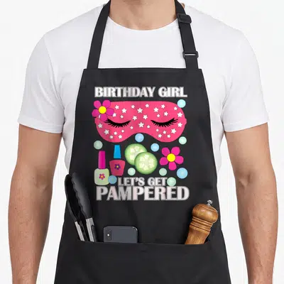 Spa birthday apron - Pink sleep mask with stars, cucumber slices, nail polish, and BIRTHDAY GIRL text — shown on a black cotton canvas apron against a white background