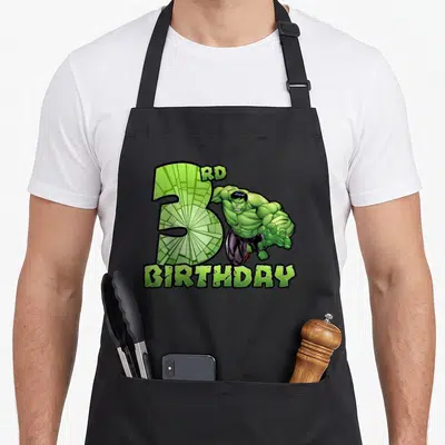 Smash 3rd birthday apron - Hulk character beside large green number 3 with RD BIRTHDAY text in cartoon style — shown on a black cotton canvas apron against a white background