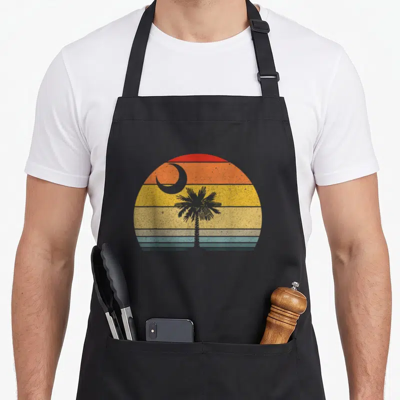 Carolina tree apron - Grey palmetto tree and crescent moon silhouetted against retro striped sunset semi-circle — shown on a black cotton canvas apron against a white background