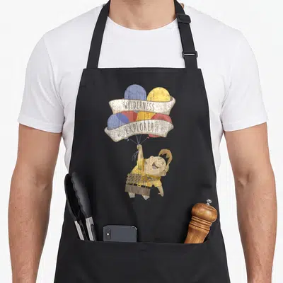 Russell balloon apron - Cartoon boy holding balloons with WILDERNESS EXPLORER banners in yellow, blue, red, and beige — shown on a black cotton canvas apron against a white background