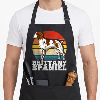 Brittany spaniel apron - Standing Brittany Spaniel dog in front of retro striped sun with stacked white text — shown on a black cotton canvas apron against a white background