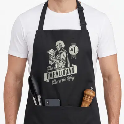 Papalorian father day apron - Distressed white print of The Mandalorian holding Grogu with Papalorian text and galaxy — shown on a black cotton canvas apron against a white background
