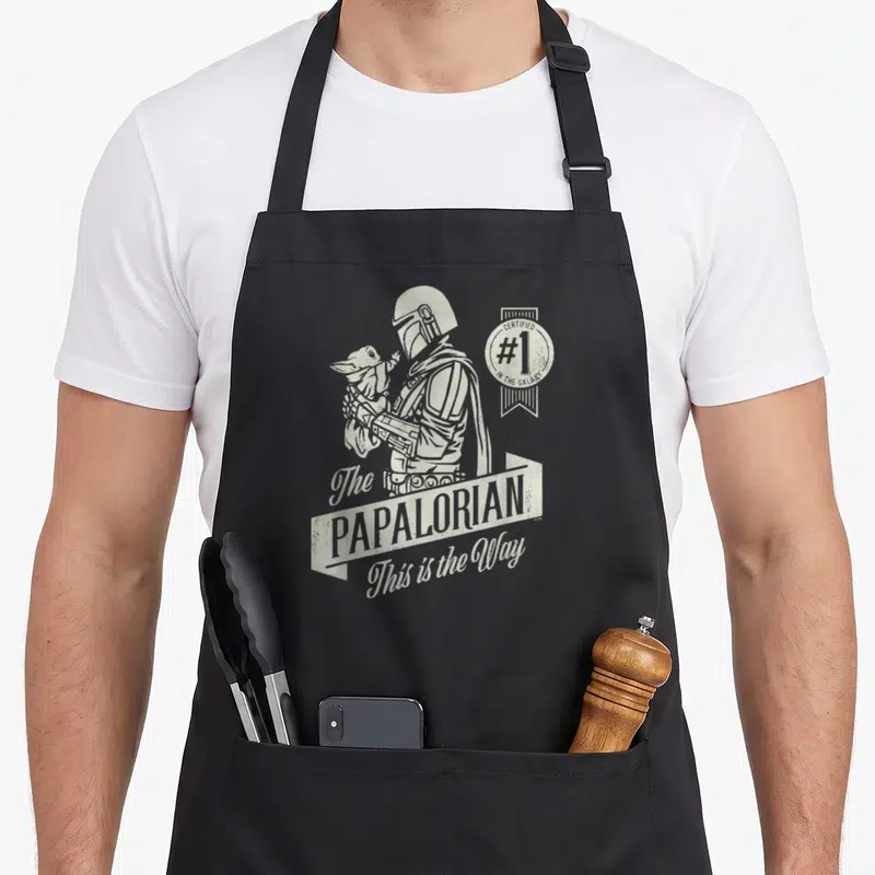 Papalorian father day apron - Distressed white print of The Mandalorian holding Grogu with Papalorian text and galaxy — shown on a black cotton canvas apron against a white background